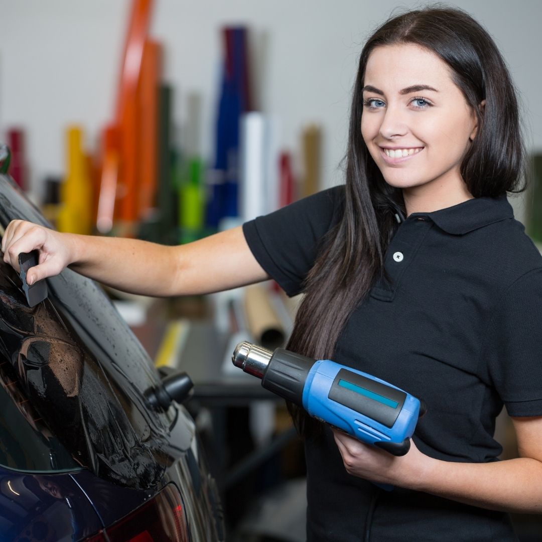 A woman smiling while using a heat gun on a vehicle wrap